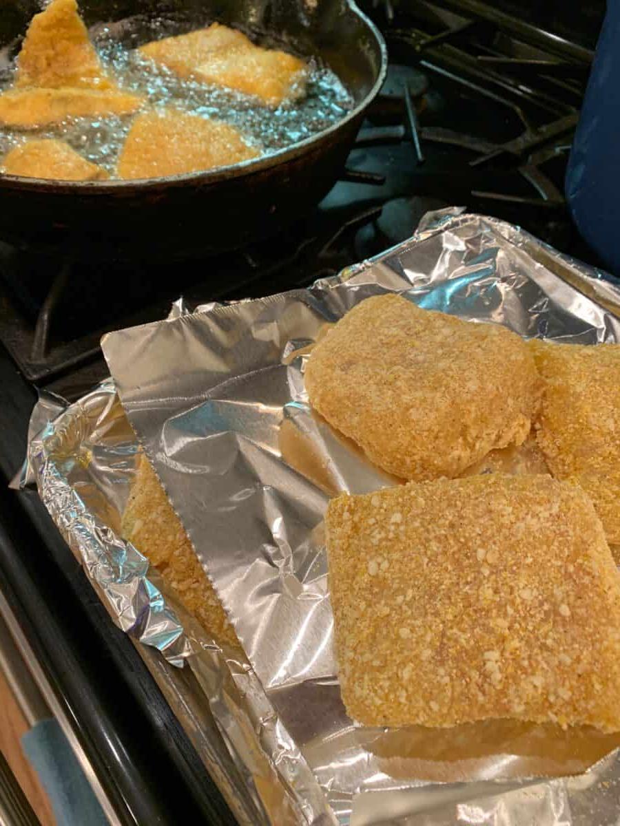Breaded cot on foil next to a skillet, and several pieces of fish cooking in the skillet.