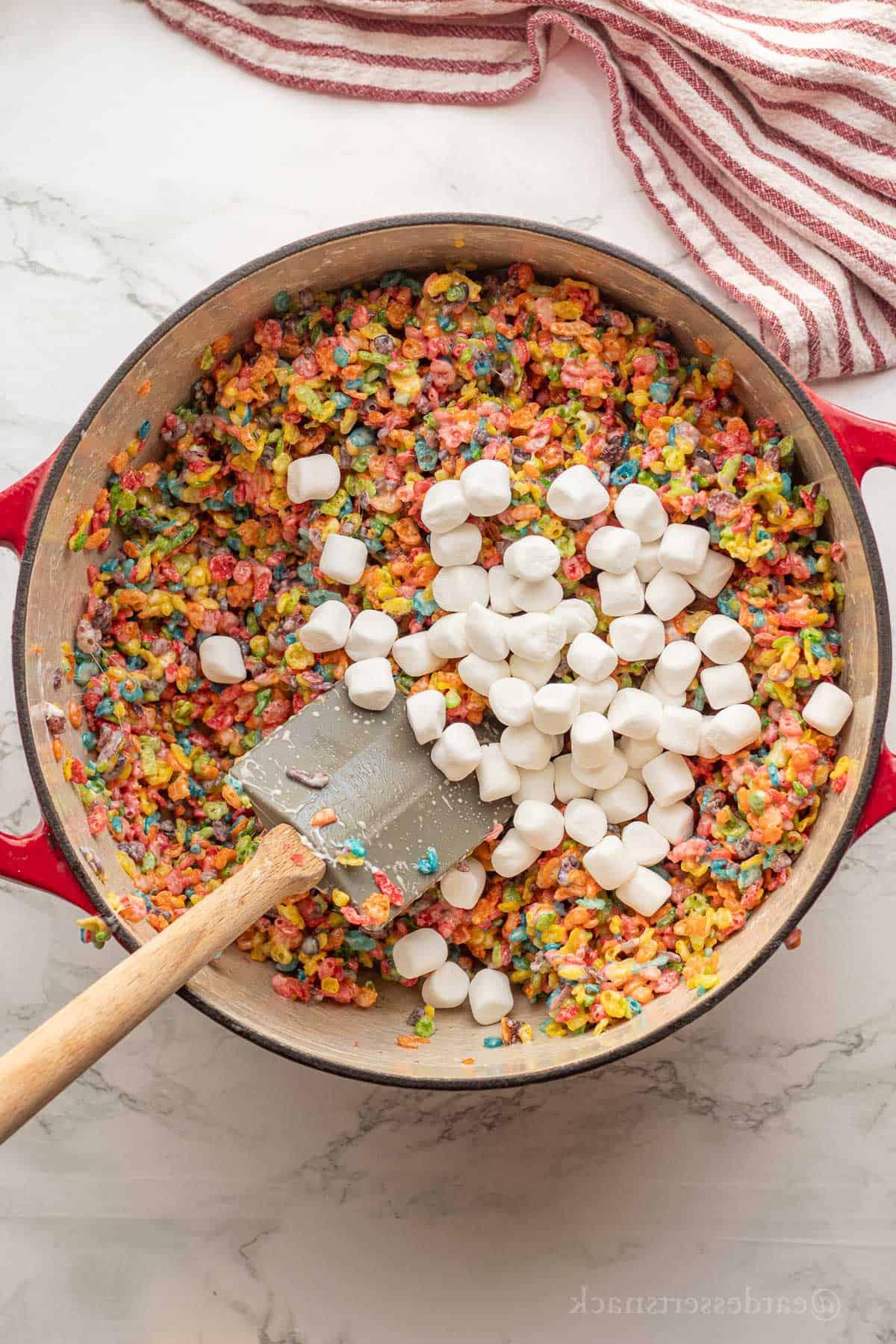 Marshallows in fruity pebble mixture in red pot with rubber spatula.