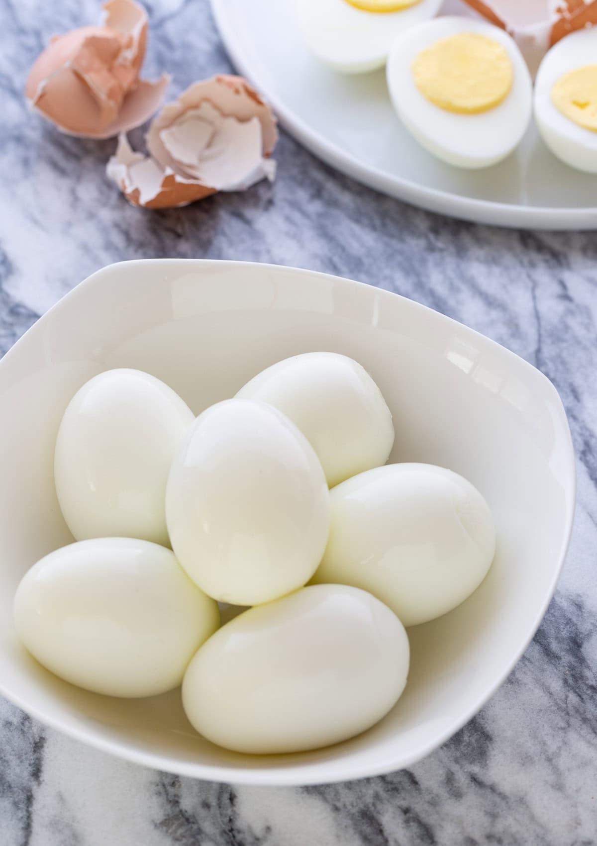 A small white bowl of peeled hard boiled eggs with egg shells in the back.